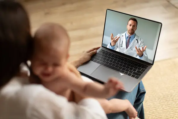 Caring mother holding crying baby and using laptop, making video call to pediatrician at home, doctor consulting online. Telemedicine and child healthcare concept