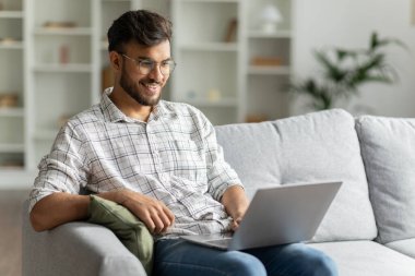 Pozitif Hintli adam ev içi koltukta dizüstü bilgisayar kullanarak çalışıyor, adam internette sörf yapıyor, modern teknolojiler kullanıyor, serbest alan kullanıyor.