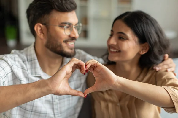 Love, affection and bonding concept. Happy indian couple making heart shape with fingers, selective focus on hands, blurred background