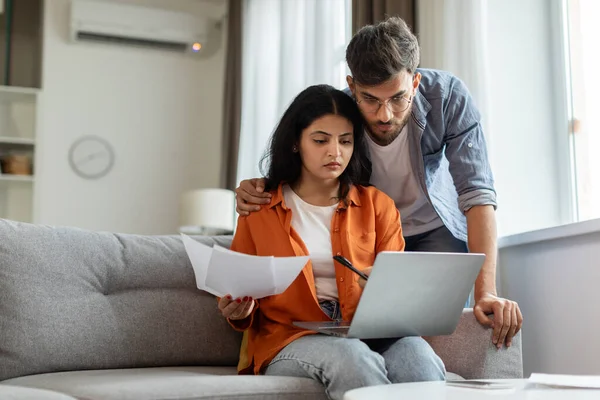 Serious indian couple paying bills online, sitting on couch, using laptop, concentrated husband and wife checking papers and banking on Internet, copy space