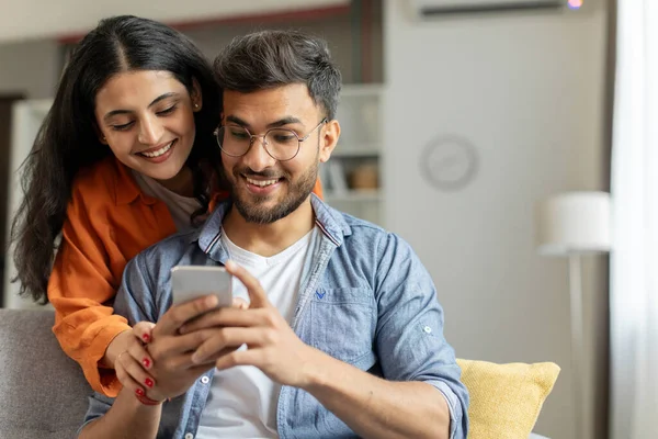 Cheerful indian couple using cellphone, shopping online or sharing social media, spouses sitting on sofa at home interior, woman embracing her husband looking at cellphone screen