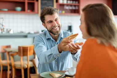 Gülümseyen Avrupalı bir adamın karısına sandviç verirken portresi, kafede otururken kadını beslemesi, boş alan. Besleyici kahvaltı, atıştırmalık