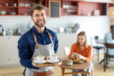 Neşeli erkek garson tepside kahve fincanları taşıyor ve kameraya gülümsüyor, küçük işletme restoranında çalışıyor.