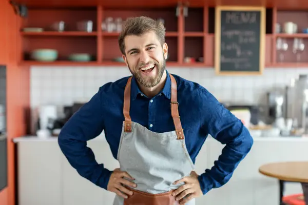 Portrait of excited handsome european male barista standing at coffee cafe counter and smiling at camera, coffee shop owner