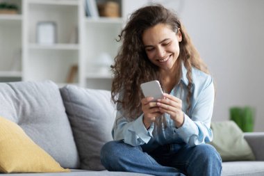 Akıllı telefon kullanan mutlu Avrupalı kadın, arkadaşlarıyla mesajlaşıyor ya da alışveriş için yeni bir uygulama arıyor, evdeki kanepede dinleniyor, uzayı kopyalıyor.