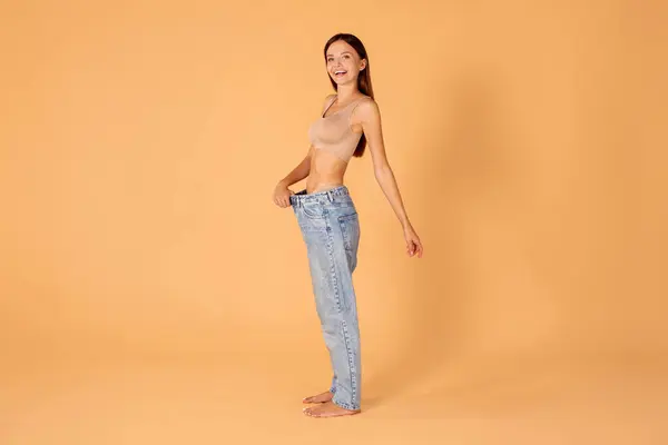 Successful weight loss diet. Lady in big jeans demonstrating results of her slimming program promoting healthy eating, side view, peach studio background