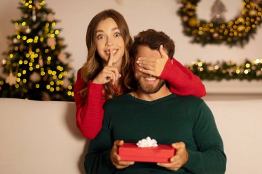 Kafkasyalı mutlu kadın sessizlik işareti gösteriyor, gözlerini kapatıyor hediye kutusunu tutuyor, arka planda Xmas ağacı ve çelenklerle kanepede oturuyor.