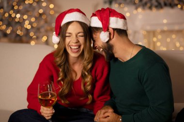 Gülen genç Avrupalı adam ve Noel Baba şapkalı bayan oturma odasında Xmas ağacı ve parıldayan çelenklerle sarılıyorlar.