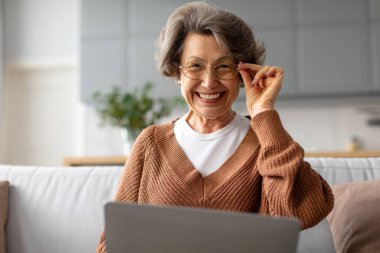 Dizüstü bilgisayar kullanan, gözlüklere dokunan, kameraya gülümseyen, evdeki kanepede oturup dijital zevkin tadını çıkaran mutlu bir son sınıf kadını.