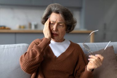 Kıdemli kadın şakaklarına masaj yapıyor, gözlük tutuyor, baş ağrısı çekiyor, evdeki kanepede oturuyor, boş yer var.