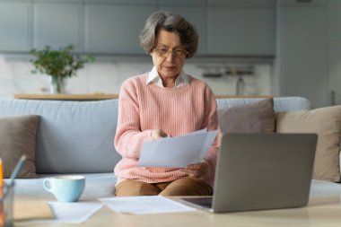 Gözlüklü ciddi bir kadın dizüstü bilgisayarla çalışıyor ve oda içi belgeler, faturalar, evden alınan vergiler.