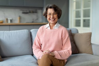 Huzurlu yaşlı bayan kameraya gülümsüyor, evde dinleniyor, oturma odasının iç kısmında koltukta oturuyor, refahın, boş alanın tadını çıkarıyor.