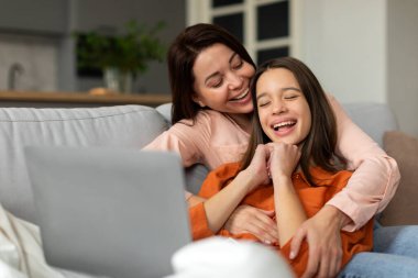 Mutlu bir kadın gülen kızına sarılırken dizüstü bilgisayar kullanıyor, internette sörf yapıyor ya da evdeki kanepede film seyrediyor.