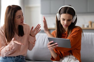 Teenage girl in headphones using digital tablet and gesturing stop to mother, while mom scolding daughter, sitting on sofa at home clipart