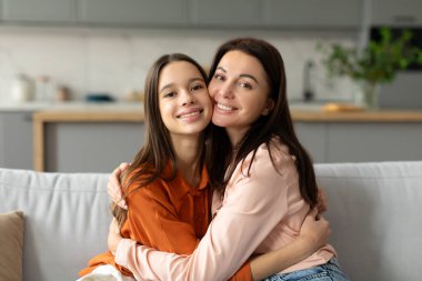 Sevgi dolu bir anne, mutlu ergen kızını kucaklıyor, ergen çocuğuna sevgisini sunuyor, birlikte kanepede oturuyor ve sarılıyor.