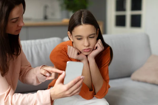 Angry young mother showing cellphone to upset teenage girl while daughter looking at screen with indifference, sitting together on sofa at home
