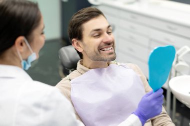 Modern klinikte diş tedaviden sonra aynaya bakan mutlu bir adam. Erkek hasta stomatoloji dolabında oturuyor ve yeni gülüşünün tadını çıkarıyor.