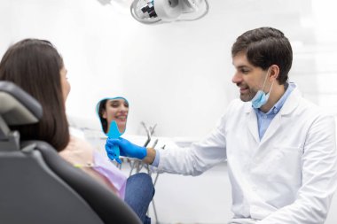 Kadın aynaya bakıyor, dişlerini kontrol ediyor klinikte diş tedavisinden sonra erkek dişçi kadın hastaya terapi sonuçlarını gösteriyor.