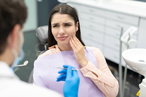 Young woman in dental chair suffering from acute tooth pain, touching cheek and looking at doctor