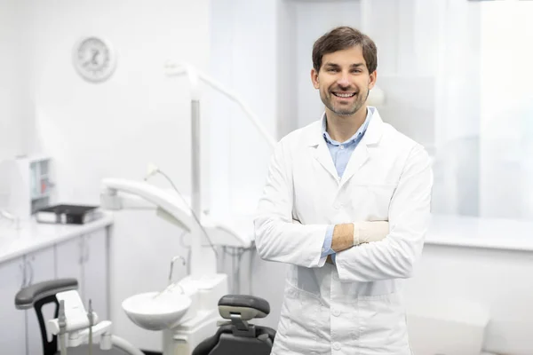 Diş Merkezi. İş yerinde poz veren erkek dişçi portresi, modern klinikte kolları bağlı duran stomatolog.