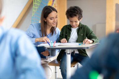 Young female teacher showing how to do task to little latin schoolboy, kids sitting at desks, studying in elementary school classroom clipart