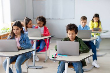 Çeşitli okul çocukları dizüstü bilgisayarlarıyla masalarında oturup dijital cihazlar kullanarak ders çalışıyorlar. Modern temel eğitim kavramı