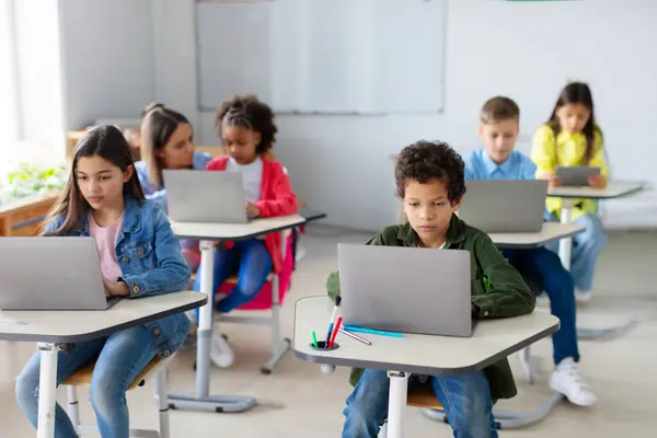 Çeşitli okul çocukları dizüstü bilgisayarlarıyla masalarında oturup dijital cihazlar kullanarak ders çalışıyorlar. Modern temel eğitim kavramı