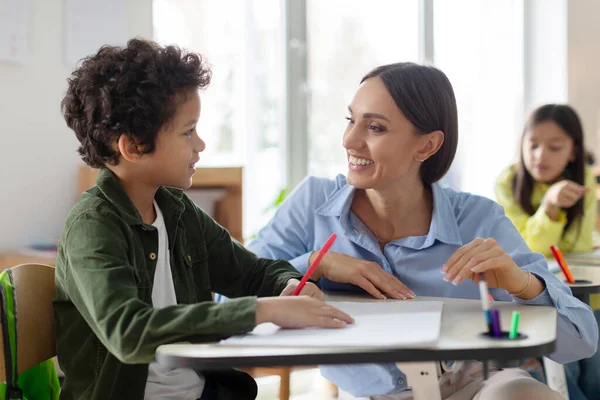 Teacher helping latin boy with assignment in elementary school, classroom support, scholarship and educator aid, explaining or helping male learner in class