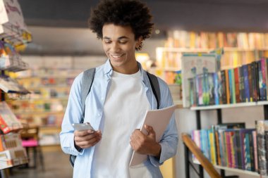 Sırt çantalı siyah erkek öğrenci kütüphanede teknik ara veriyor, elinde cep telefonu, denge çalışması ve dijital bağlantı kullanıyor.