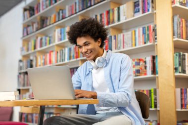 Kütüphanede online ders için dizüstü bilgisayar kullanan siyahi öğrenci sessiz çalışma ortamında eğitime modern yaklaşımı sergiliyor.