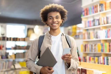 Kütüphanede elinde dijital tablet ve cep telefonu olan mutlu siyah erkek öğrenci modern teknolojiyle geleneksel çalışma yöntemlerini birleştiriyor.