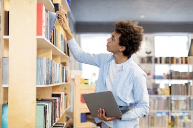 Siyah erkek öğrenci, elinde dizüstü bilgisayarla kütüphanedeki raflardan kitap alıyor. Teknolojinin ve geleneksel kitap alımının dinamik karışımının örneğini veriyor.