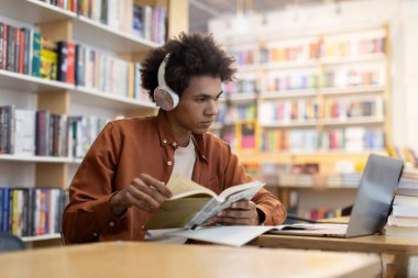 Kulaklıklı siyahi öğrenci dizüstü bilgisayara bakıyor ve elinde kitap tutuyor. Üniversite kütüphanesinde okuyor. Yoğunlaştırılmış odaklanma ve harmanlanmış öğrenme yöntemleri var.