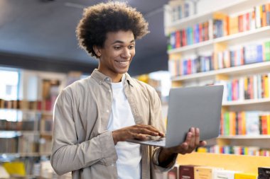 Afrikalı Amerikalı öğrenci dizüstü bilgisayar kullanıyor ve gülümsüyor, kütüphanenin içinde duruyor, teknolojinin pozitif kesişimini yakalıyor ve öğreniyor.