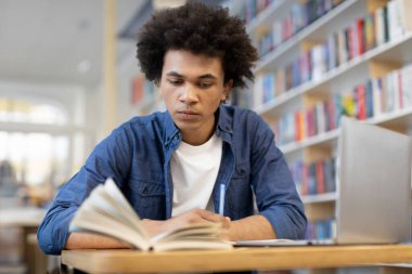 Odaklanmış siyah erkek öğrenci kitap okuyor ve not alıyor, üniversite kütüphanesinde dizüstü bilgisayarın önünde oturuyor, geleneksel okuma ve teknolojiyle geliştirilmiş not almanın dinamik kombinasyonu