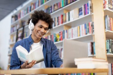 Kütüphanede dizüstü bilgisayarın önünde oturan, kulaklık takan, kitap tutan ve bilgisayar ekranına bakan siyahi öğrenci.