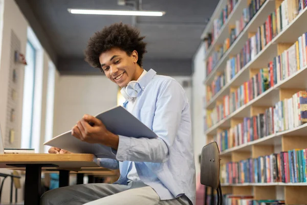 Smiling black male student sitting in the library, writing notes in copybook, holding book, radiating cheerfulness in cozy library, free space