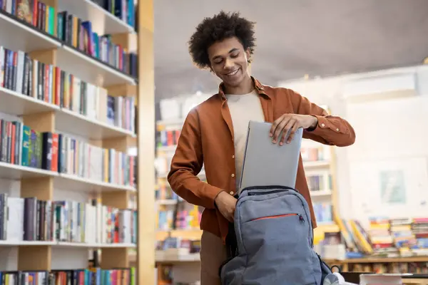 Smiling black student guy packing laptop in backpack in university library, moment capturing the conclusion of successful study session