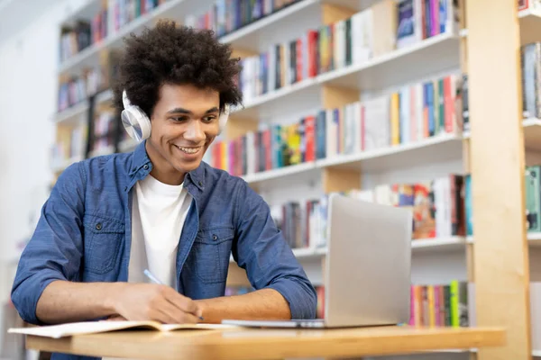 Focused black student guy in headphones taking notes and looking at laptop screen while sitting in university library, scene of concentration and technology-enhanced learning