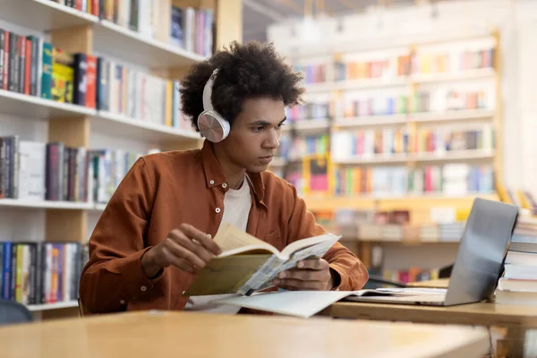 Focused black student guy in headphones looking at laptop screen and holding copybook, studying in university library, scene of concentrated focus and blended learning methods