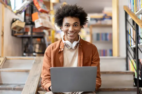 Happy black student guy with headphones on neck sitting on stairs in university library, using laptop and smiling at camera