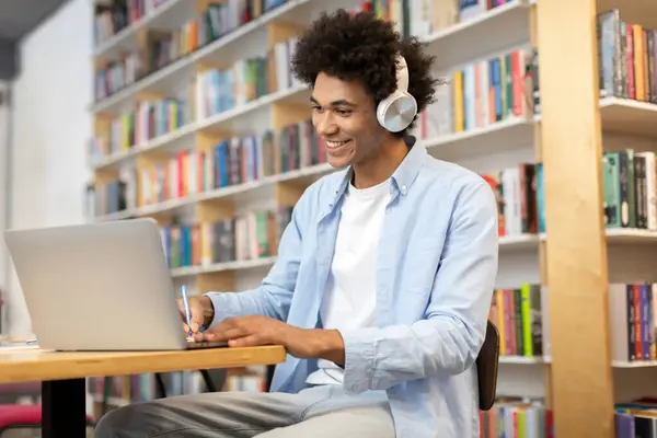 Black student guy sitting in library, wearing headphones, using laptop and diligently writing notes, creating dedicated and immersive study session