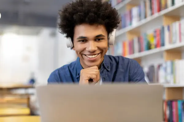 Black student guy, wearing headphones, looking at laptop screen with smile, sitting in library, perfect blend of focused study and cheerful technology use