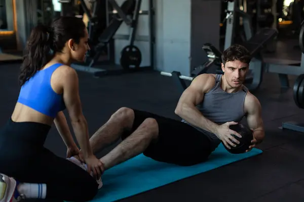 Woman trainer helping man training with ball, doing abs workout, guy doing exercise on press with trainer, modern gym interior