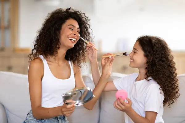 Joyful girl applying makeup on her moms face, sitting on sofa at home, having family spa day, putting on different cosmetics, enjoying weekend together