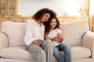 Mutlu anne ve küçük kız akıllı telefon kullanıyor evdeki koltukta oturuyor, sosyal medyada kayıyor, telefon aygıtıyla web sörfü yapıyor.