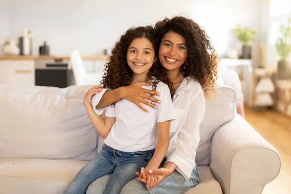 Bonding concept. Portrait of happy mom and daughter hugging and smiling at camera, enjoying time together, sitting on sofa at home
