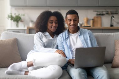 Romantik siyah eşler evdeki rahat koltukta dizüstü bilgisayarla dinleniyor, çift bilgisayar kullanıyor, internette geziniyor ya da alışveriş yapıyor.