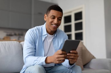 Yakışıklı siyahi adam evde dijital tablet kullanıyor, modern aletlerle koltukta dinleniyor, eğlence için teknolojilerin tadını çıkarıyor, yer kopyalıyor.