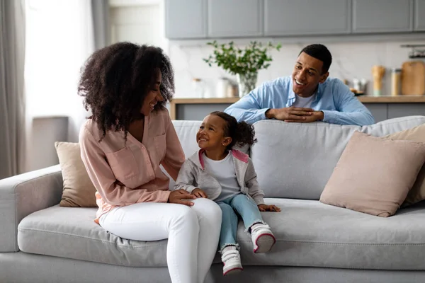 Happy black parents resting with cute little daughter, sitting on sofa in living room interior, loving family of three posing at home